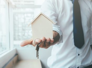 Hand agent with home in palm and key on finger. Offer of purchase house, rental of Real Estate. Giving, offering, demonstration, handing house keys.