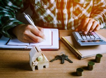 Selective focus wood home key and step up gold coin or currency on wooden table with document.Blur hand man calculate finance and writing on notebook at home office desk.business concept background.